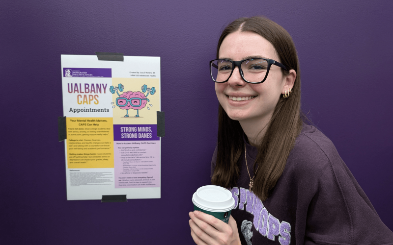 Izzy D'Ambro stands next to a small poster display of her social media messages for making an appointment at UAlbany's Counseling Center. She holds a cup of coffee.