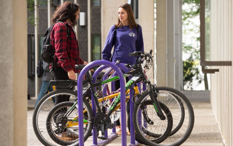 Two women talk while standing next to bikes in a purple bike rack.