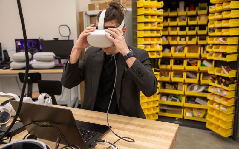 A researcher puts on a virtual reality headset, while seated at a table with a laptop.