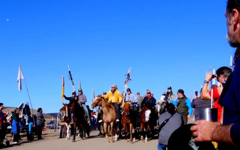 A group of indigenous protesters stand, some riding horses, protesting against the construction of the Dakota Access Pipeline.