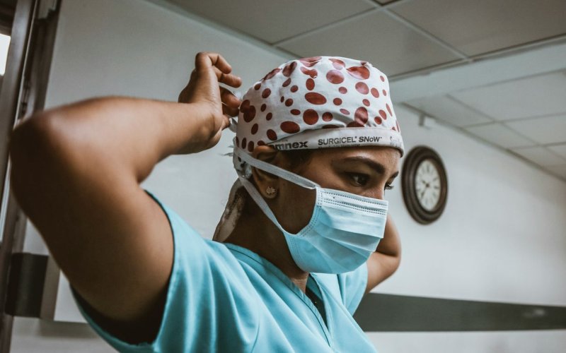 A person wearing light blue medical scrubs and a surgical cap secures a paper face mask around their head. 