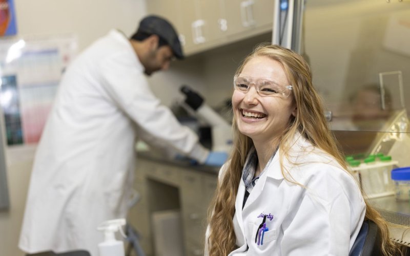 A woman with long wavy blond hair, wearing a white lab coat and clear protective goggles, smiles for a portrait in a lab. Lab benches with test tubes, and a man looking into a microscope, are in the background.