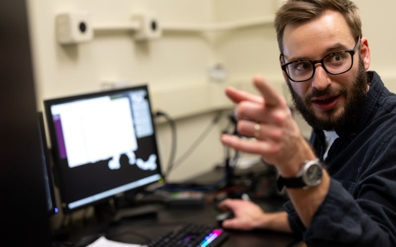 A man with eyeglasses sits in front of a computer screen turned toward the camera and pointing.