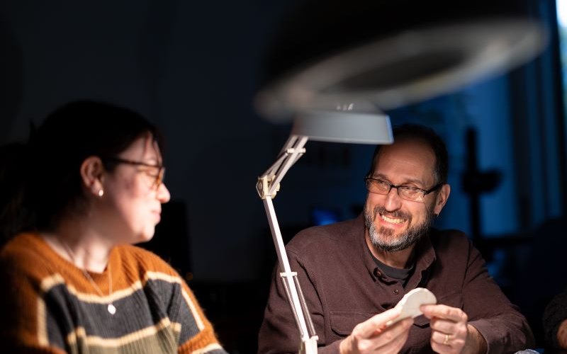 A man smiling and seated at a table holds a piece of pottery illuminated by a desk lamp while talking to a woman seated next to him.