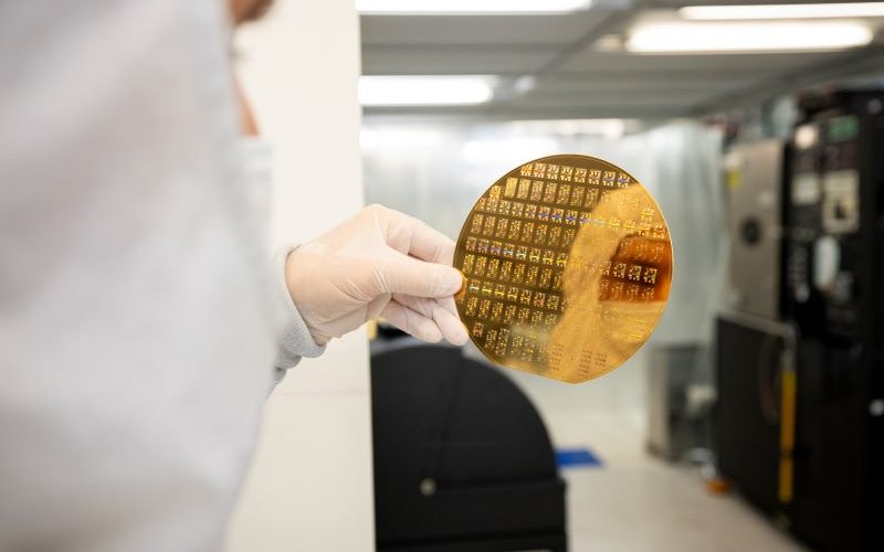 A gloved hand holds a gold-colored silicon wafer reflecting the image of a person in a cleanroom "bunny" suit.