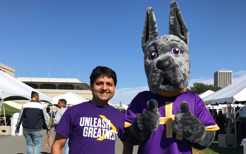 Khem Sedhai, wearing a purple t-shirt with the UAlbany slogan, "Unleash Greatness," smiles and poses for a photo with the UAlbany mascot, Damien the Great Dane.