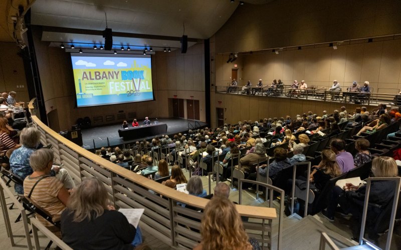 In a large auditorium people in stadium-style seating listen to a panel discussion on a stage at the bottom with a large visual display behind the panelists reads Albany Book Festival