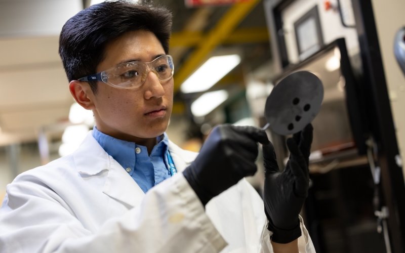 A man with short black hair in safety goggles, white lab coat and black gloves inspects a piece of lab equipment.