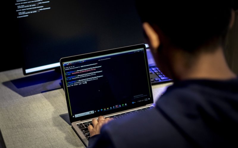 A man sits at a desk with a desktop computer and types code on a laptop.