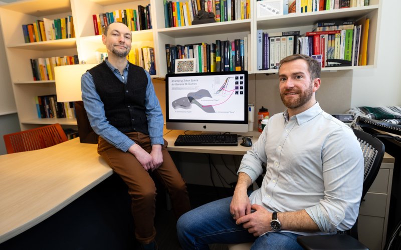 Two professors, one sitting on a desk with a black vest and blue collared shirt, the other seated at a computer, look facing the camera as a graphic display of AI in gaming is shown on an Apple iMac computer with a row of books behind the computer.