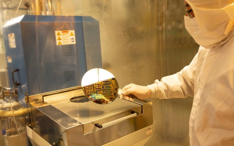 A man in a cleanroom "bunny suit" holds up a 200mm silicon wafer used for microchip fabrication.