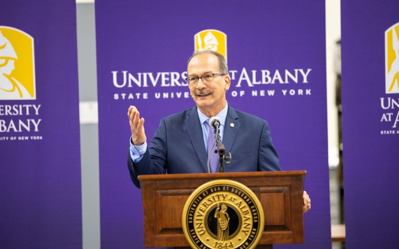 President Rodriguez stands behind a podium in front of UAlbany banners