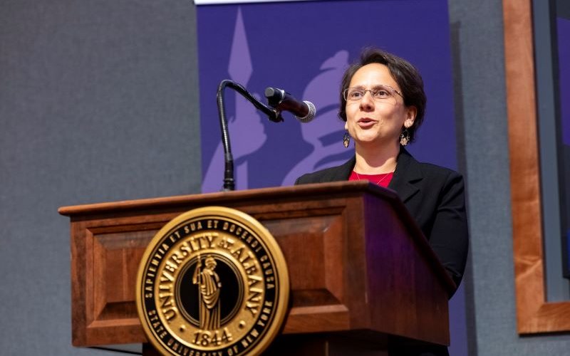 A woman in a black blazer stands behind a brown lectern with a gold University at Albany seal on the front, speaking into a microphone.