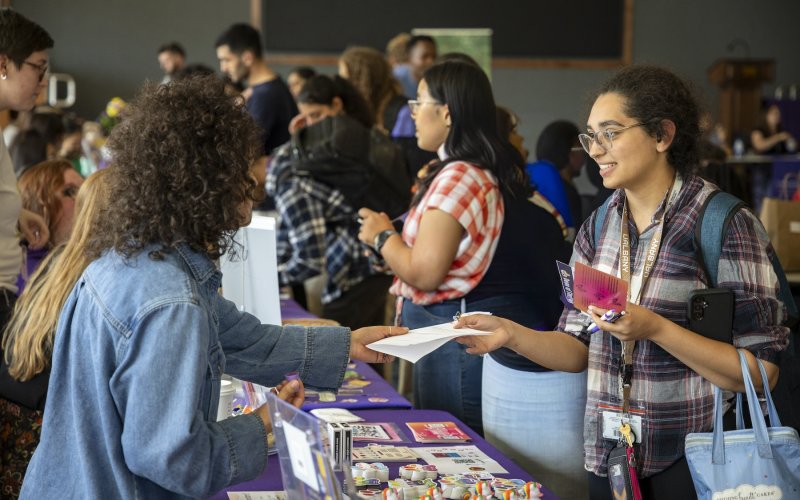 Two people interact at a busy indoor event table, exchanging papers, with informational materials and other attendees visible in the background.
