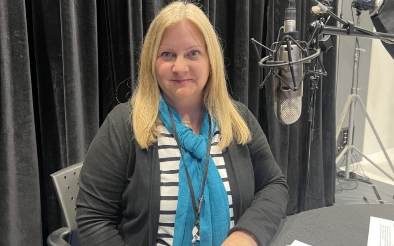 A woman with a blue scarf and a gray sweater sits at a table with a microphone hanging nearby