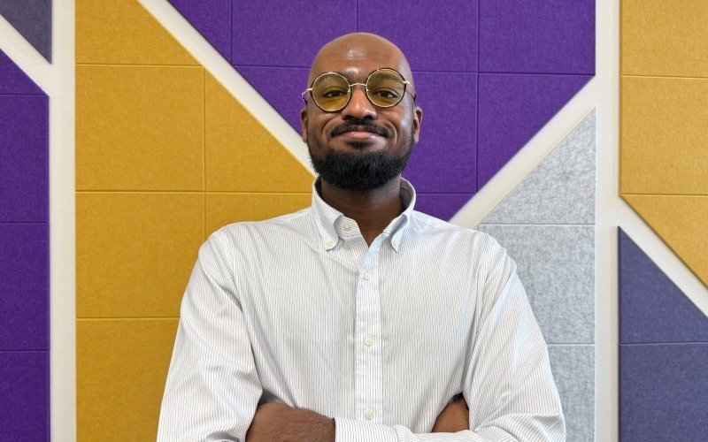 A man wearing yellow tinted glasses and a white, pinstriped button-down shirt poses for a portrait, hands folded across his chest, in front of a wall covered with decorative gold, purple and gray panels.