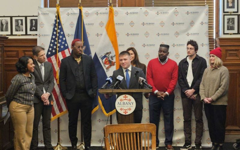 A young man in a suit speaks at a lecturn with a round plaque reading ALBANY on it, as 7 others gather around watching him