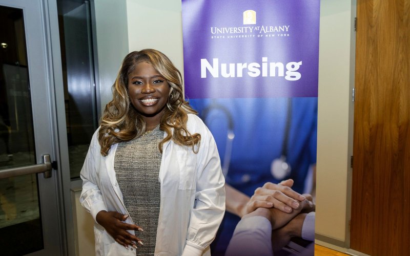 A nursing student stands in front of a poster that says "Nursing".