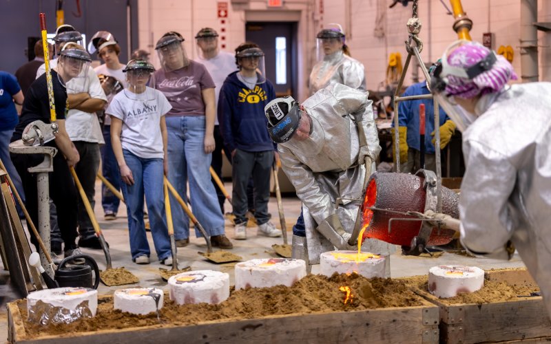Two people clad in shiny metallic protective outfits pour molten metal into a mold while a group of students observe from a safe distance.