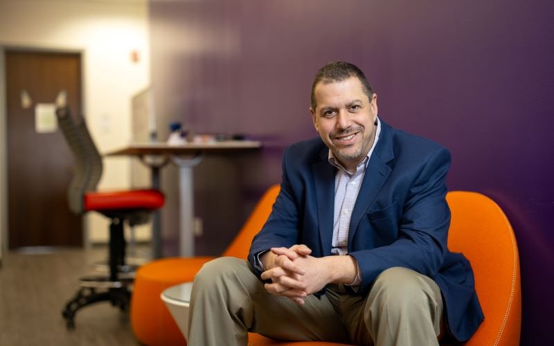 A smiling man in a blue business jacket sits smiling in an orange chair with his hands clasped. 