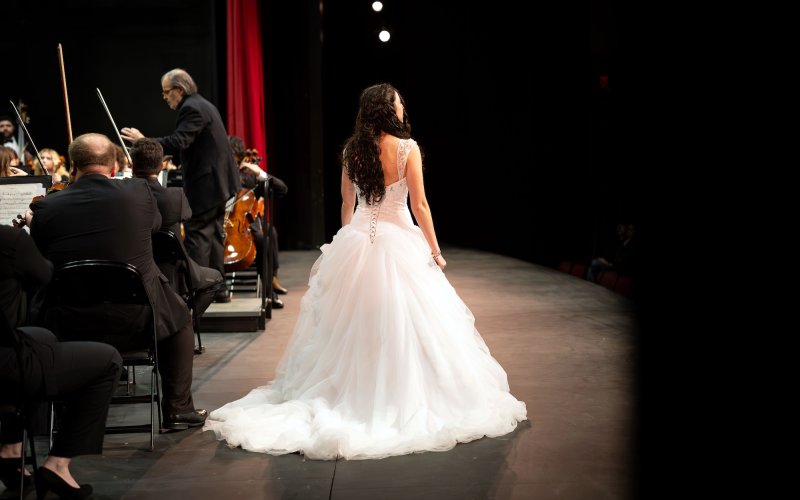 a woman in a white dress stands on stage with members of the orchestra behind her