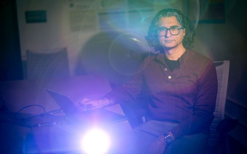 A man wearing a maroon shirt and glasses sits in a dark classroom lit by a projector creating ethereal orbs of light across the image.