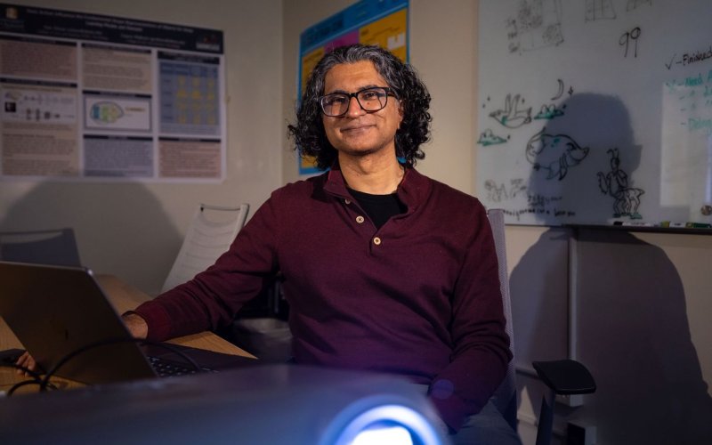 A man wearing a maroon shirt and black-rimmed glasses sits in a dark classroom lit by a projector. On the walls behind him are two colorful posters and a whiteboard containing a collection of drawings and notes. The man has dark hair and is smiling. 