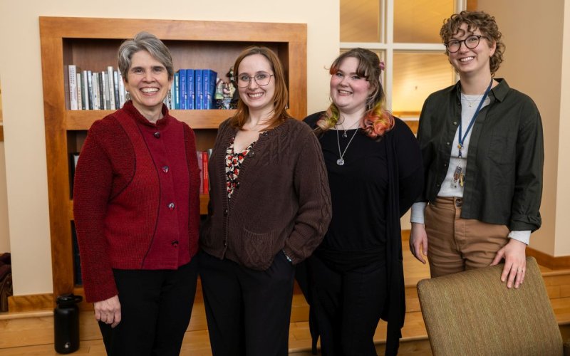 Four smiling people pose for a group portrait. They are standing in front of a bookshelf in a warmly lit room with a window behind them.