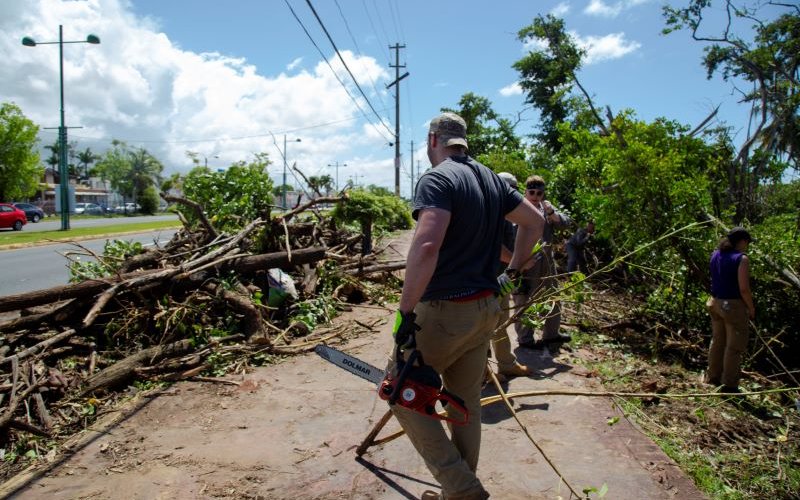 A man with his back to the camera holds a chainsaw in one hand while standing next to a pile of fallen limbs and other debris on a bright, sunny day.