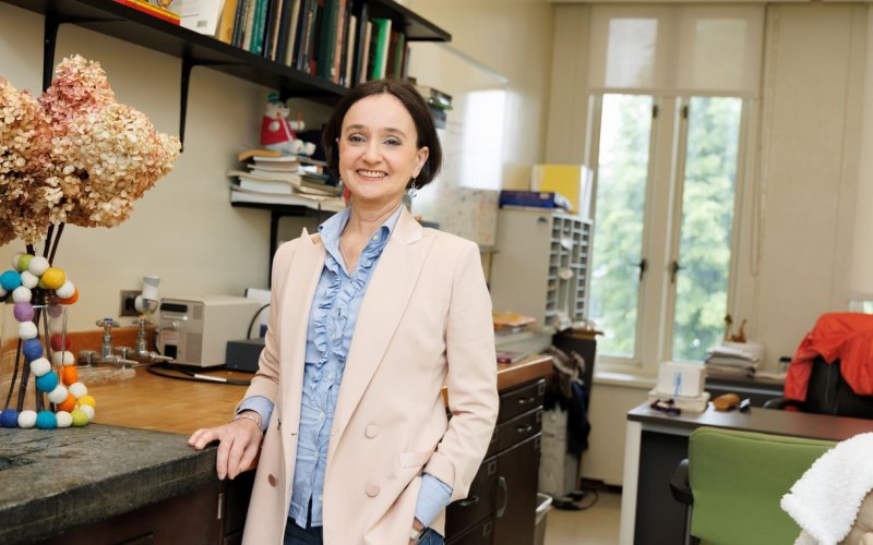 A woman with short brown hair wearing a pale pink blazer and ruffled powder-blue blouse smiles for a portrait in her office.
