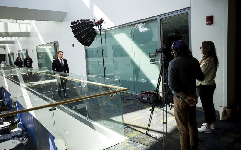 A behind-the-scenes moment shows Oscar Umana, a Massry School of Business student at the University at Albany, being interviewed. Video gear including cameras and lights are pointed at Umana as he speaks.
