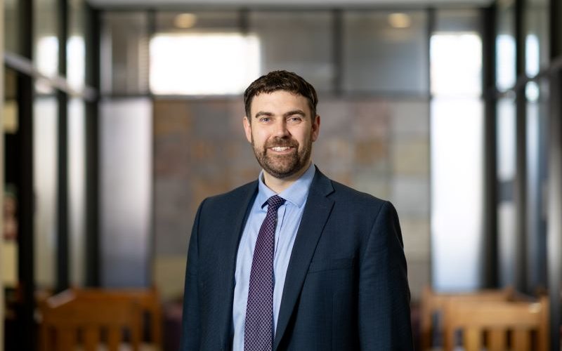 A smiling portrait of a bearded man with a dark blue suit and purple tie. 
