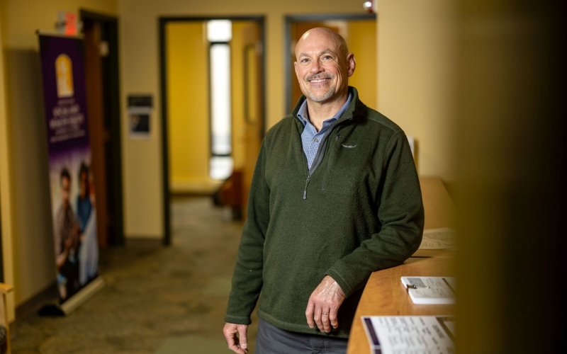 A man wearing a green quarter-zip sweater smiles for a portrait in a warmly lit hall.
