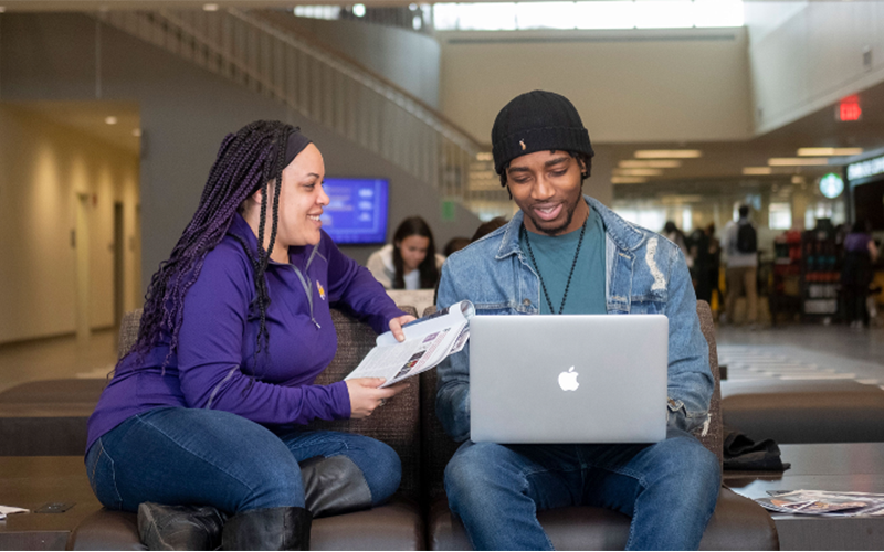Twi student sit in Campus Center, talking to each other about what they're looking at on a laptop and some paper.