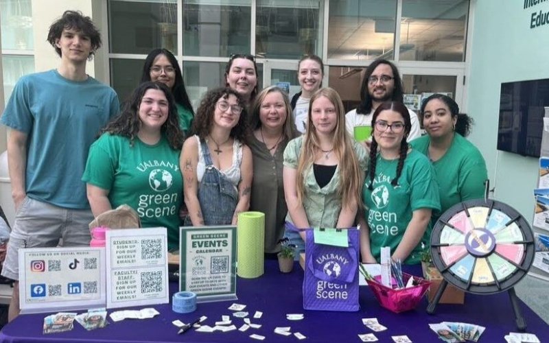 A group of people, many in green T-shirts reading UAlbany Green Scene, stand in front of a table with signs about environmental events.