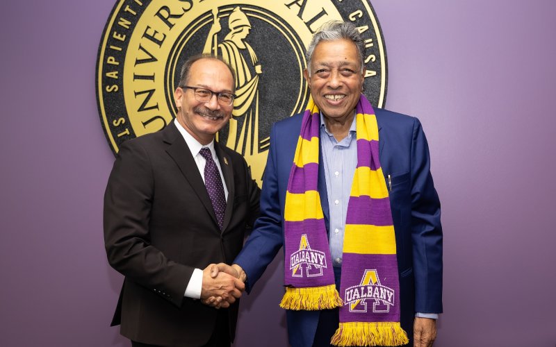 A smiling man in a blue blazer, gold and purple UAlbany scarf shakes hands with a smiling man in a black suit and purple tie.