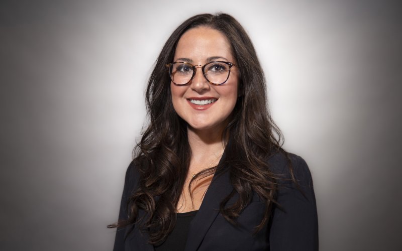 A woman with long dark hair and glasses in a black blazer smiles for a portrait against a gray backdrop.