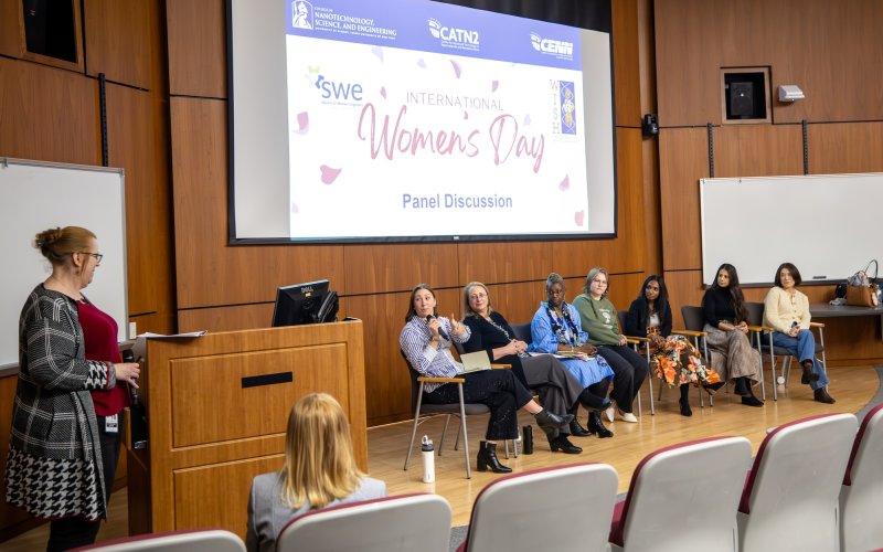 Seven women are seated on a stage as another woman asks questions from a lectern. A screen behind them says "International Women's Day Panel Discussion.""