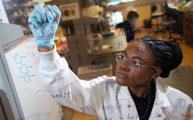 A young African American scientist wearing glasses and a white lab coat writes formulas on a clear panel with a medical lab in the background.