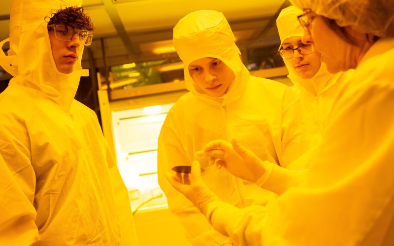 Three students in protective bunny suits examine a semiconductor wafer held by an expert.