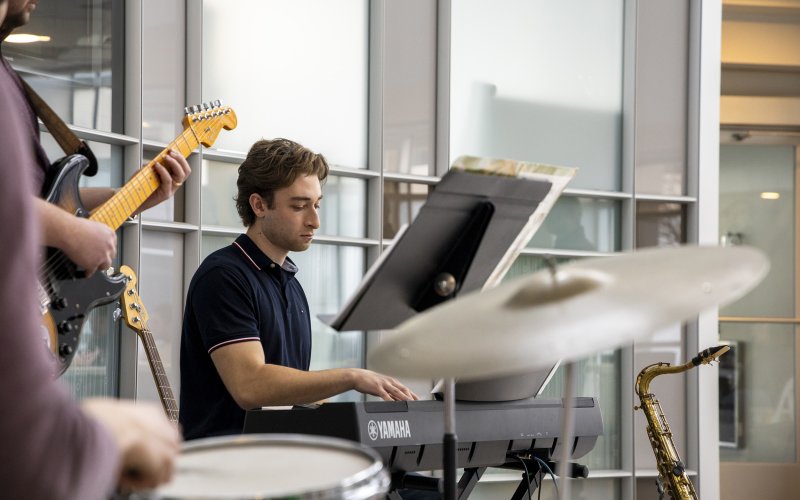 A student sits at a keyboard while musicians play drums and guitar with a cymbal in the foreground.