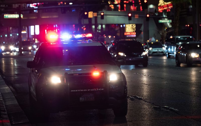 A police car with lights on is seen on a busy, wet street at night. The dark street is lit up with headlights and their reflections.