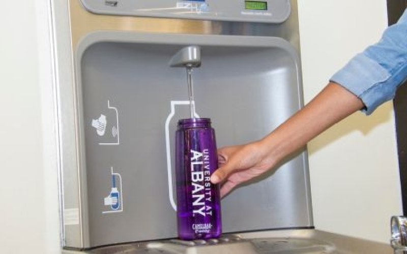 An arm and hand are seen refilling a water bottle with the words University at Albany printed on it