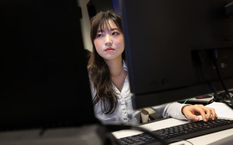 A young woman with dark hair wearing a collared button-down blue and white shirt looks at a pair of monitors in the UAlbany AI for Business Lab