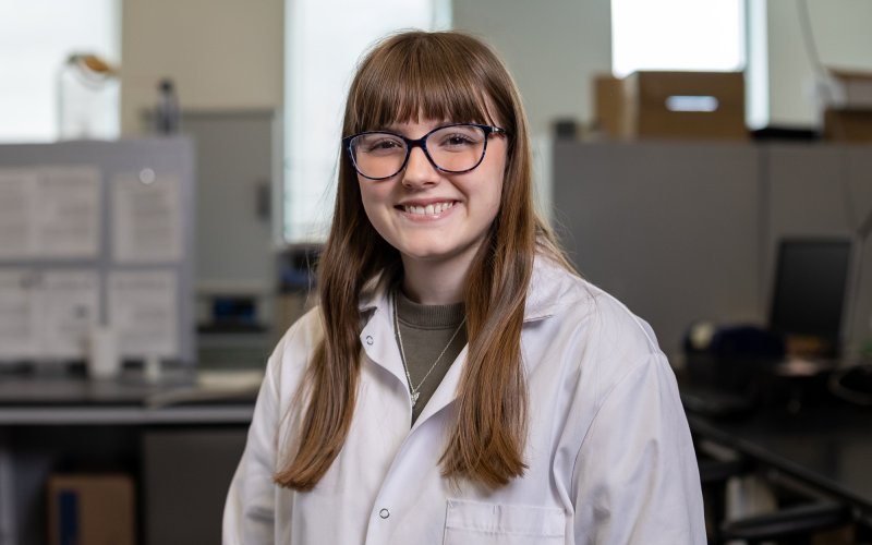 UAlbany undergraduate researcher Katelyn Jacques smiles while wearing a lab coat inside UAlbany's Paleoclimate Lab.