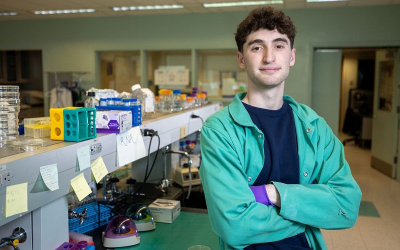 A young man with dark hair wearing a green lab coat and purple latex gloves poses for a portrait in front of a lab bench in a chemistry lab. The bench is full of various chemistry supplies including glass containers, plastic pipette stands and PostIt notes.