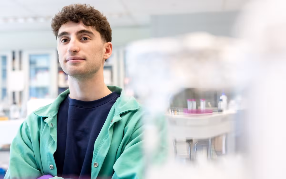 A young man with dark hair wearing a green lab coat stands in a brightly lit lab. The background is artfully blurred. 