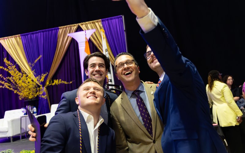 Four men dressed in suits huddle together to take a selfie with a cell phone. Three of the people are UAlbany students and one is Vice President Mike Christakis.