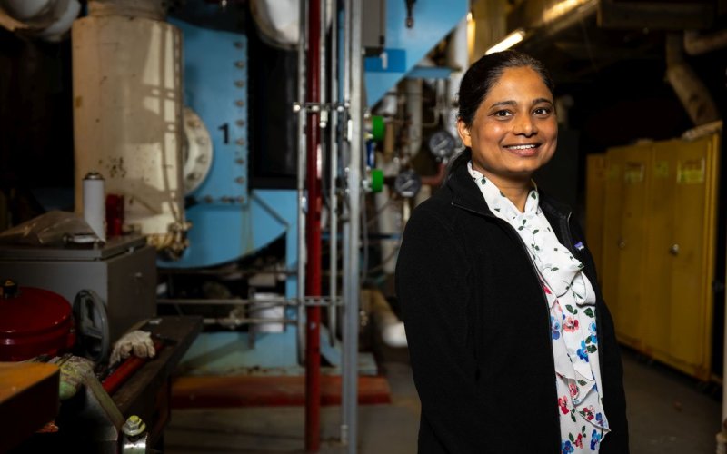 A woman wearing a black blazer and floral blouse smiles for a portrait in a dimly lit industrial room with metal equipment and lockers in the background.