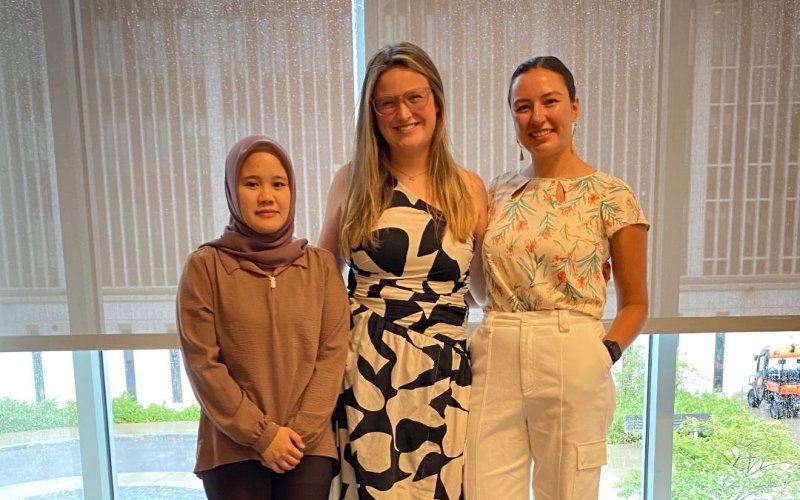 Three women stand smiling indoors by large windows. The first wears a hijab and brown outfit, the second a black and white dress, and the third a floral top and white pants. The atmosphere is friendly and bright.